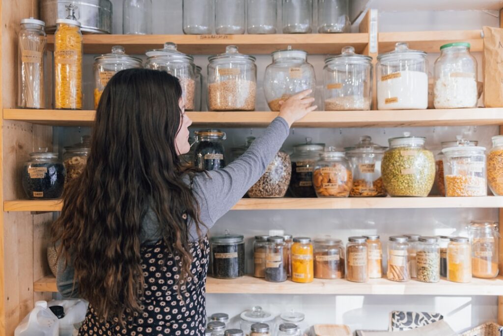 Woman with long wavy hair reaching for a glass jar on a wooden pantry shelf filled with jars of grains and spices.