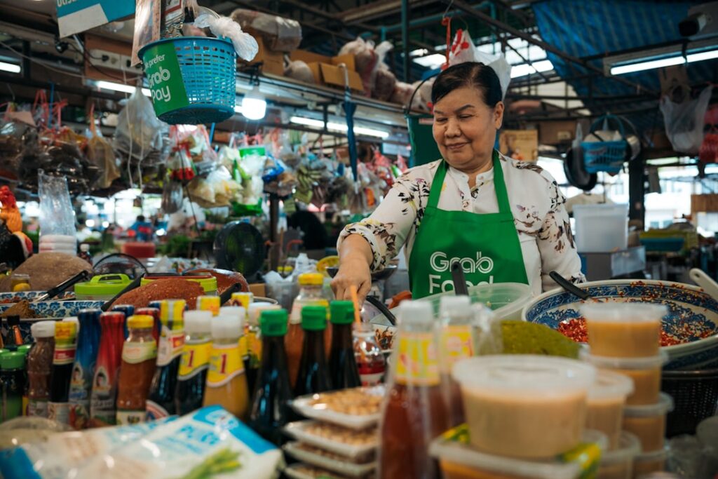 Vendor wearing a green GrabFood apron prepares sauces at a busy market stall with colorful bottles in the foreground.