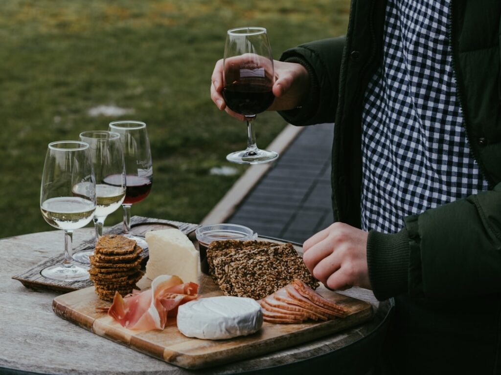 Person wearing a green jacket and checkered shirt holds a glass of red wine over a wooden board with cheese, crackers, bread, and sliced meat outdoors.