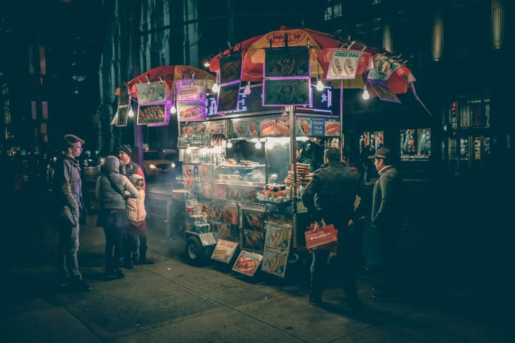Nighttime street food cart glowing with lights as customers gather around the menu.