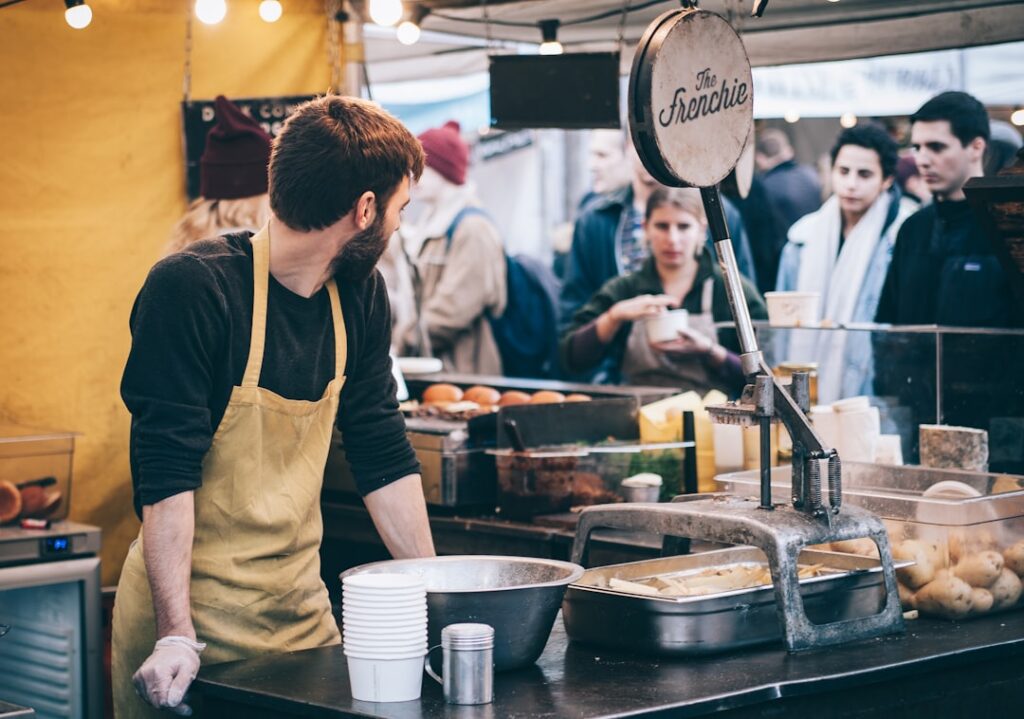 Bearded vendor in a beige apron works at an outdoor market stall, cups stacked beside him, and customers in line.