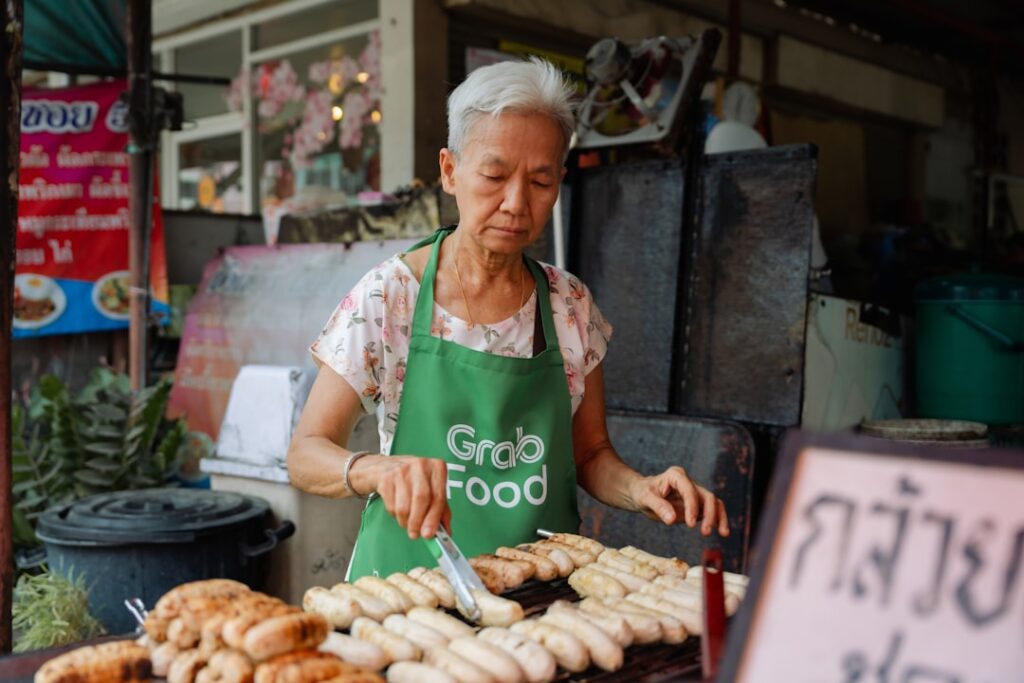 Elderly woman wearing a green GrabFood apron grills skewers at a street food stall.
