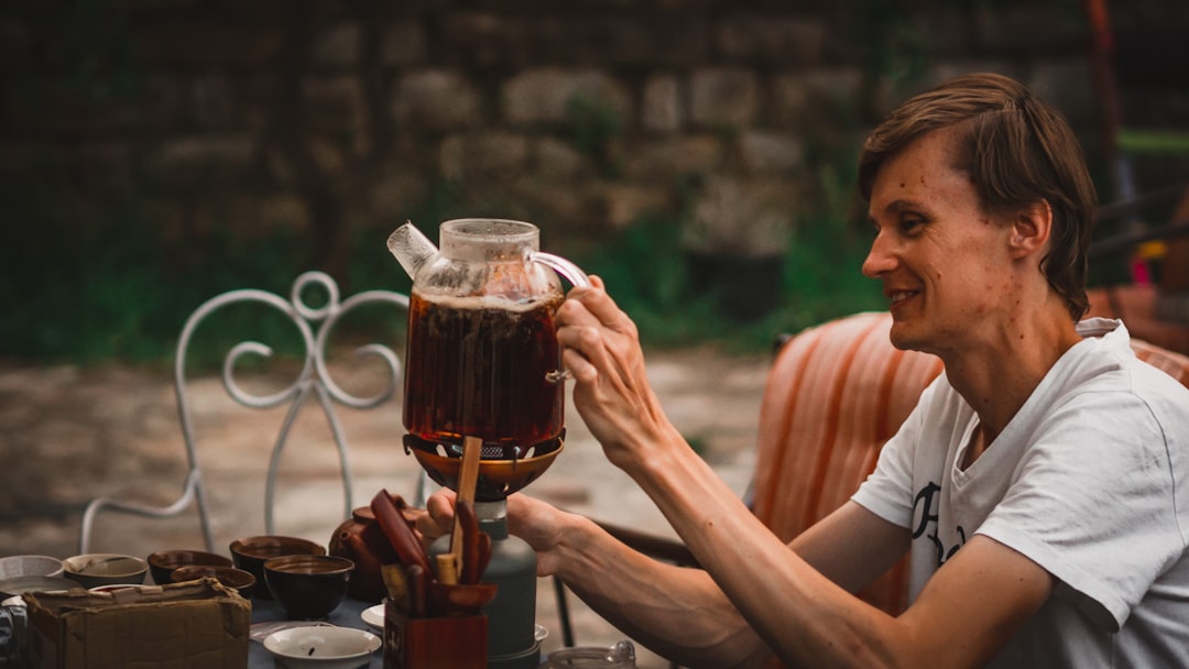 Uma mulher servindo cuidadosamente chá frio de infusão lenta de um pote de vidro em uma xícara sobre uma bancada de cozinha de madeira