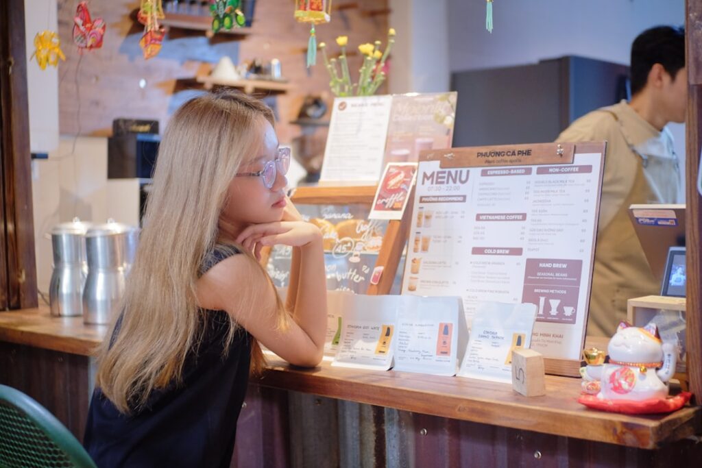 Woman with long blonde hair and glasses leaning on a cafe counter, studying a large menu board.