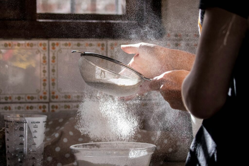 Person sifts flour through a metal sieve into a bowl in a kitchen, flour drifting in the air.