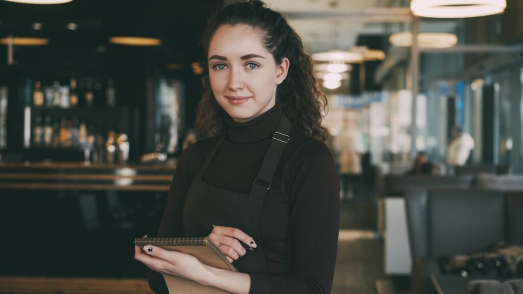 Barista in a black apron and turtleneck stands in a modern cafe, holding a notebook and smiling at the camera.