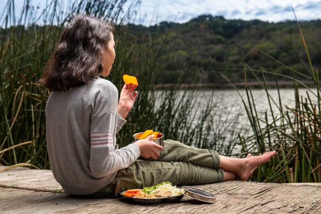 Girl sitting barefoot on a wooden dock by a lake, eating orange slices from a metal cup with a plate of food beside her.
